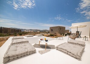 A modern outdoor seating area with cushioned sofas, a small table with a drink, and a stone building in the background. - Campo Suites (Paros)
