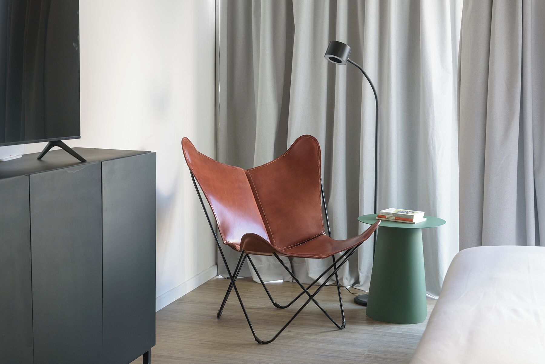 Una habitación de hotel moderna con un sillón de cuero, una mesita de noche con libros, una lámpara verde y un televisor sobre un mueble.