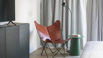 Una habitación de hotel moderna con un sillón de cuero, una mesita de noche con libros, una lámpara verde y un televisor sobre un mueble.