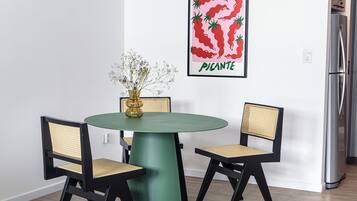 Un comedor con una mesa verde, dos sillas negras con asientos de mimbre, un jarrón con flores y un cuadro en la pared con forma de chile picante y la palabra “PICANTE”.