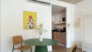 Un coin repas moderne avec une table verte, deux chaises en bois et un vase contenant des fleurs séchées. Un tableau abstrait aux couleurs vives est accroché au mur.