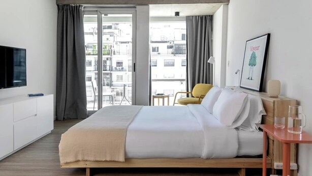 Une chambre d’hôtel moderne dotée d’un grand lit, d’une télévision, d’une petite table avec une lampe et offrant une vue sur la ville grâce à de grandes fenêtres.