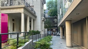 Un étroit sentier piétonnier bordé de plantes en pot, une structure rose et un bâtiment doté de colonnes et d’un balcon.