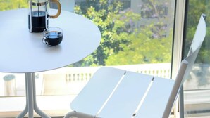 Une table ronde avec une machine à café et une tasse, une chaise blanche, et une vue sur un bâtiment à travers une fenêtre en verre.