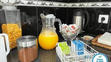 A kitchen counter with a coffee maker, a glass pitcher, a container of cereal, a container of coffee, and a basket of tea bags.