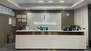 Un comptoir d’accueil orné du logo de l’hôtel Urban Garden, une horloge et un écran numérique.