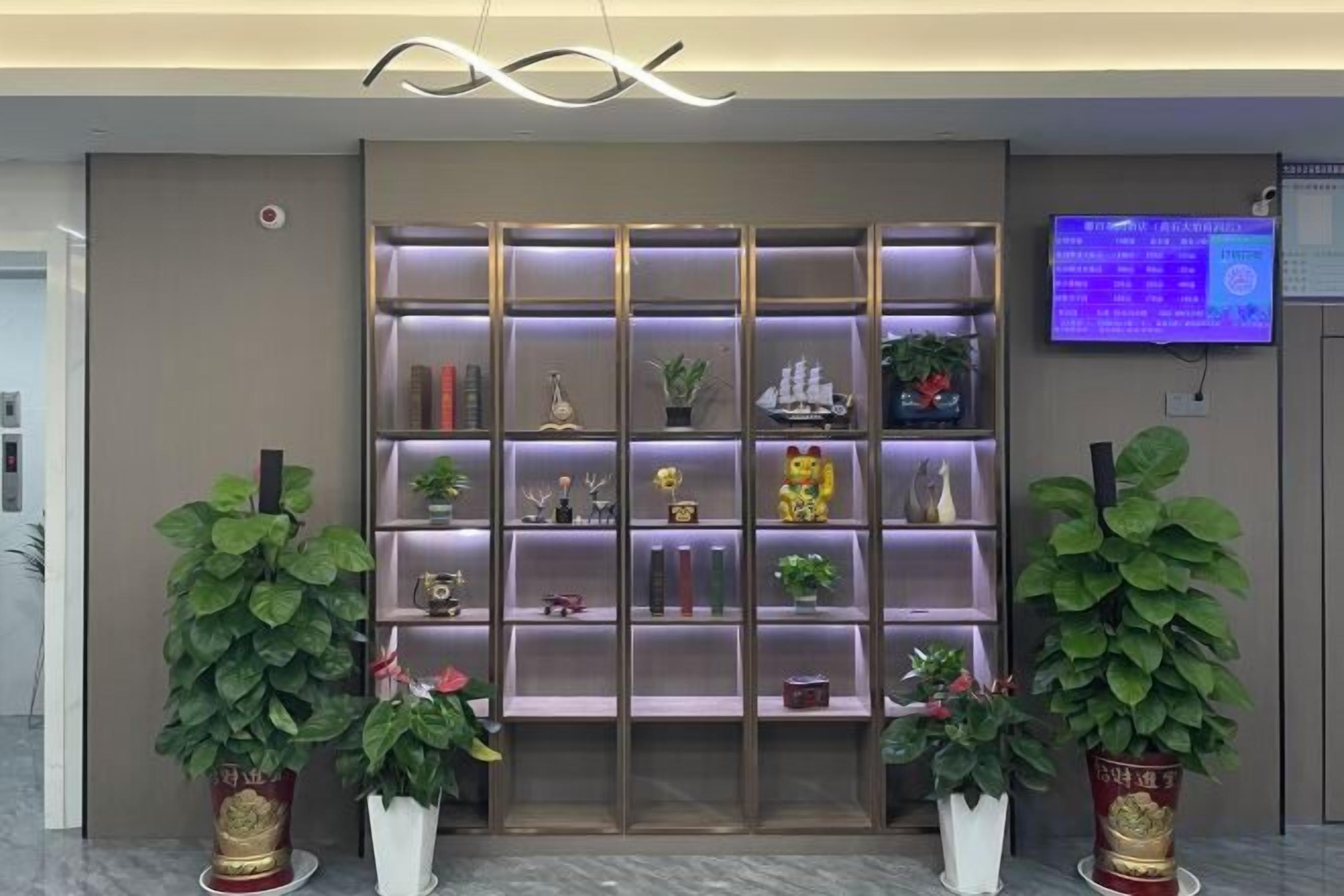 Un área de recepción con una estantería moderna que exhibe diversos objetos decorativos y plantas, un panel informativo digital y dos plantas en macetas.
