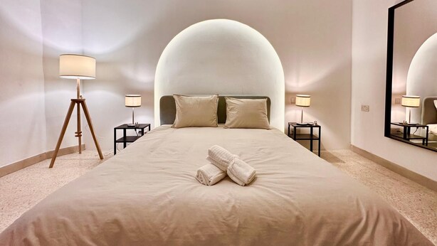 A modern hotel room with a large bed, two bedside tables with lamps, a large mirror, and a unique curved ceiling design.