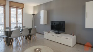 A modern living room with a dining area, featuring a flat-screen TV, white cabinetry, and a glass-top dining table with chairs. - Apt24 - The Roof in Desenzano del Garda (Desenzano del Garda)