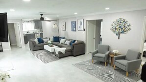 Un salon moderne avec un canapé d’angle gris, une table basse blanche et un fauteuil gris. On y trouve un tapis à motifs noir et blanc, un ventilateur de plafond et un élément décoratif mural.