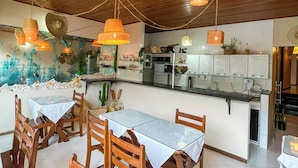 A dining area with wooden chairs and tables, a kitchen in the background, and hanging wicker lamps. - Pousada Estevão (Cairu)