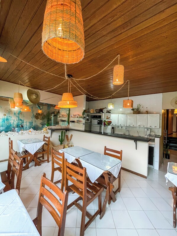 A dining area with wooden chairs and tables, a kitchen in the background, and hanging wicker lamps.