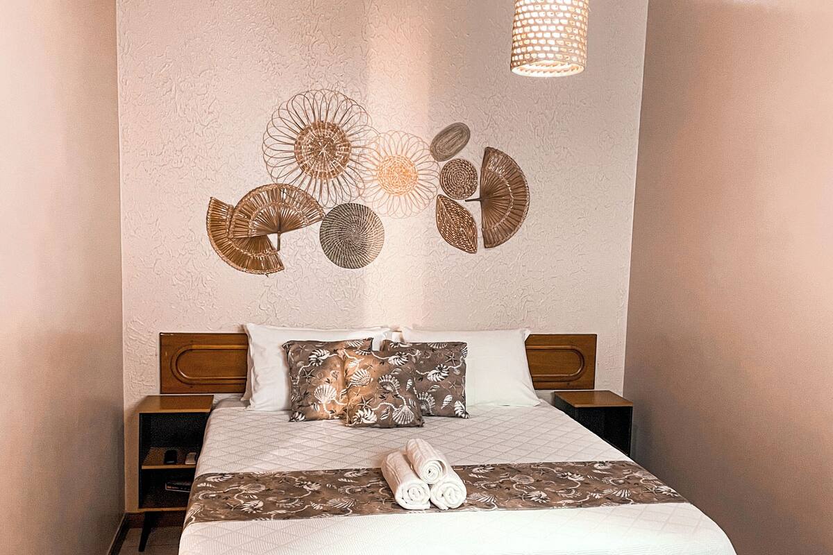 Un lit bien fait, avec une couette blanche et des coussins décoratifs marron, un luminaire suspendu et une œuvre d’art murale au-dessus du lit.