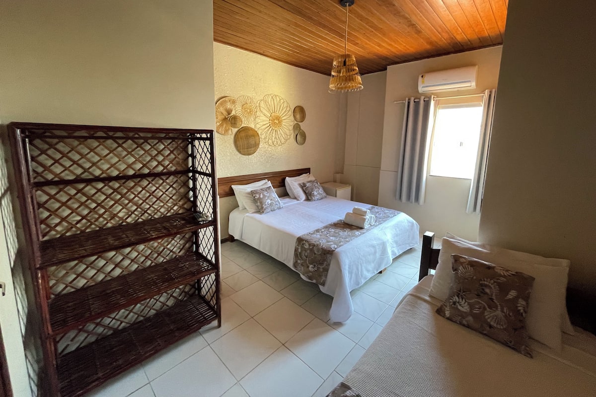 Une chambre à coucher avec une tête de lit en bois, un lit recouvert de draps blancs, un luminaire suspendu et une fenêtre avec des rideaux.