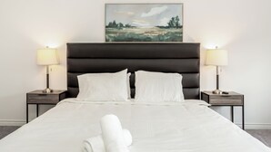 Un lit bien fait, avec du linge de lit blanc et des serviettes pliées, flanqué de deux tables de chevet avec des lampes, dans une pièce où un grand tableau est accroché au-dessus du lit.