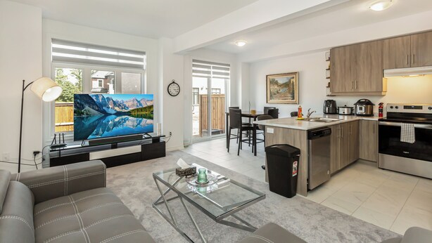 Un salon moderne avec un téléviseur à écran plat, une table basse en verre et un canapé. La cuisine est équipée d’un lave-vaisselle, d’une cuisinière et de meubles de rangement en bois.