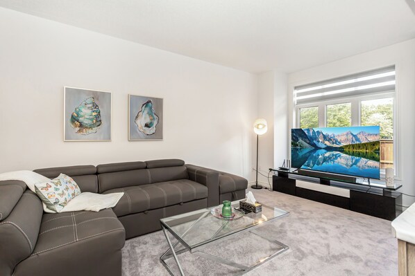 Una sala de estar moderna con un sofá modular, una mesa de centro de vidrio y un televisor de pantalla plana. Se observan dos obras de arte enmarcadas en la pared y una lámpara de pie.