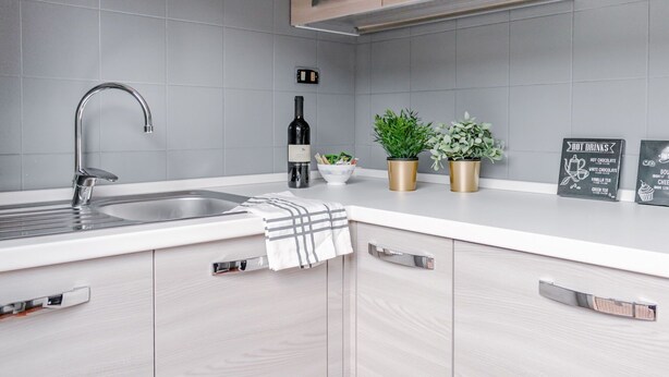 Cozinha moderna com azulejos cinza, pia de aço inoxidável, armários de madeira e bancada com uma garrafa de vinho, plantas em vasos e um cardápio em lousa.