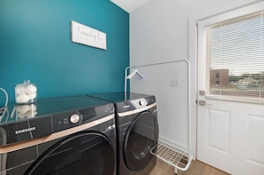 Un área de lavandería con dos lavadoras Samsung, una mesa de vidrio con un tarro de algodón, un perchero y una puerta con ventana.