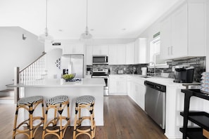 Una cocina moderna con armarios blancos, una isla central con taburetes de bar, electrodomésticos de acero inoxidable y una escalera a la vista.