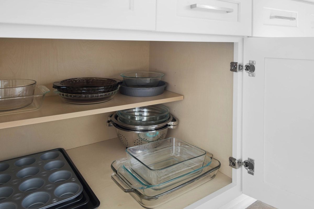A kitchen with white cabinets, a wooden shelf, and various kitchenware including a colander, bowls, and a baking tray.