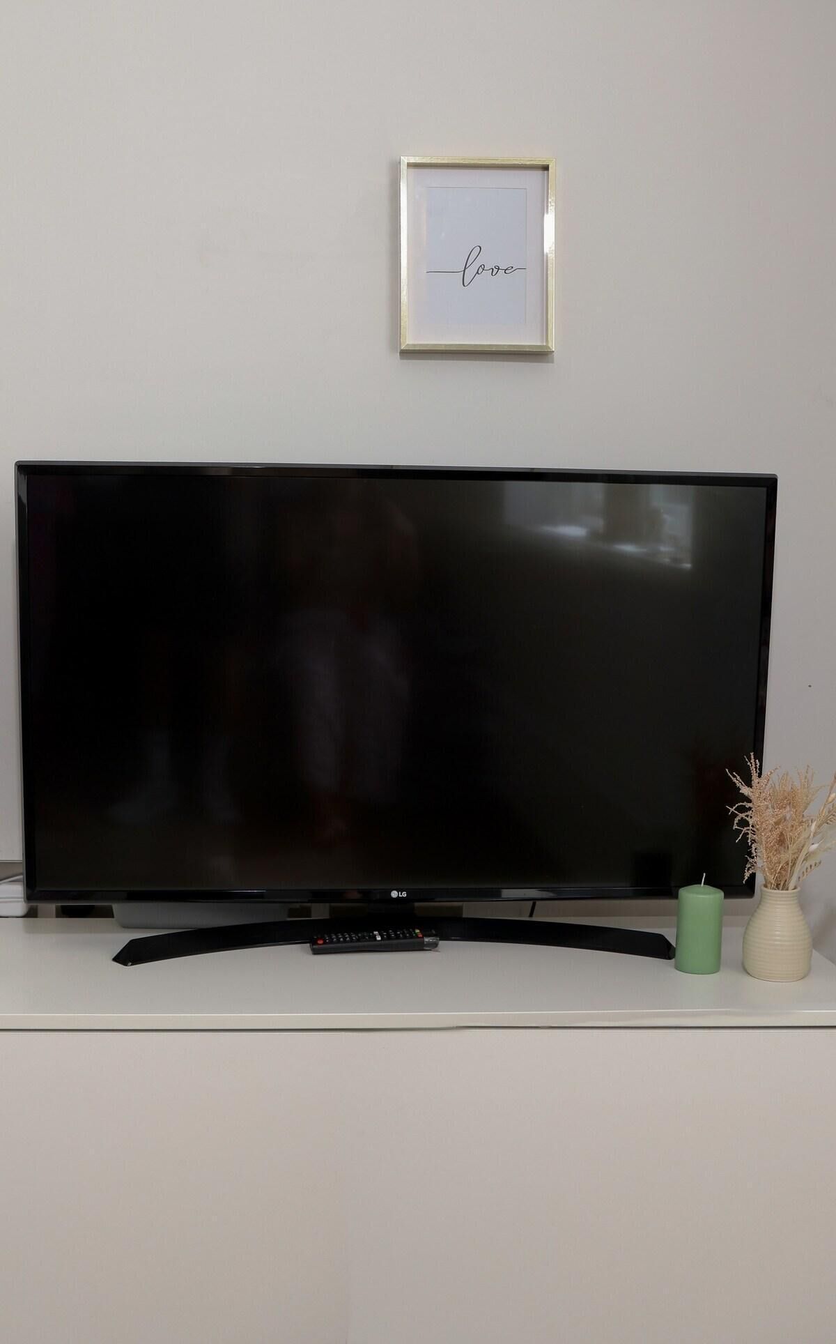 A flat-screen TV on a white stand with a remote control, a framed picture on the wall, and two vases on the stand.