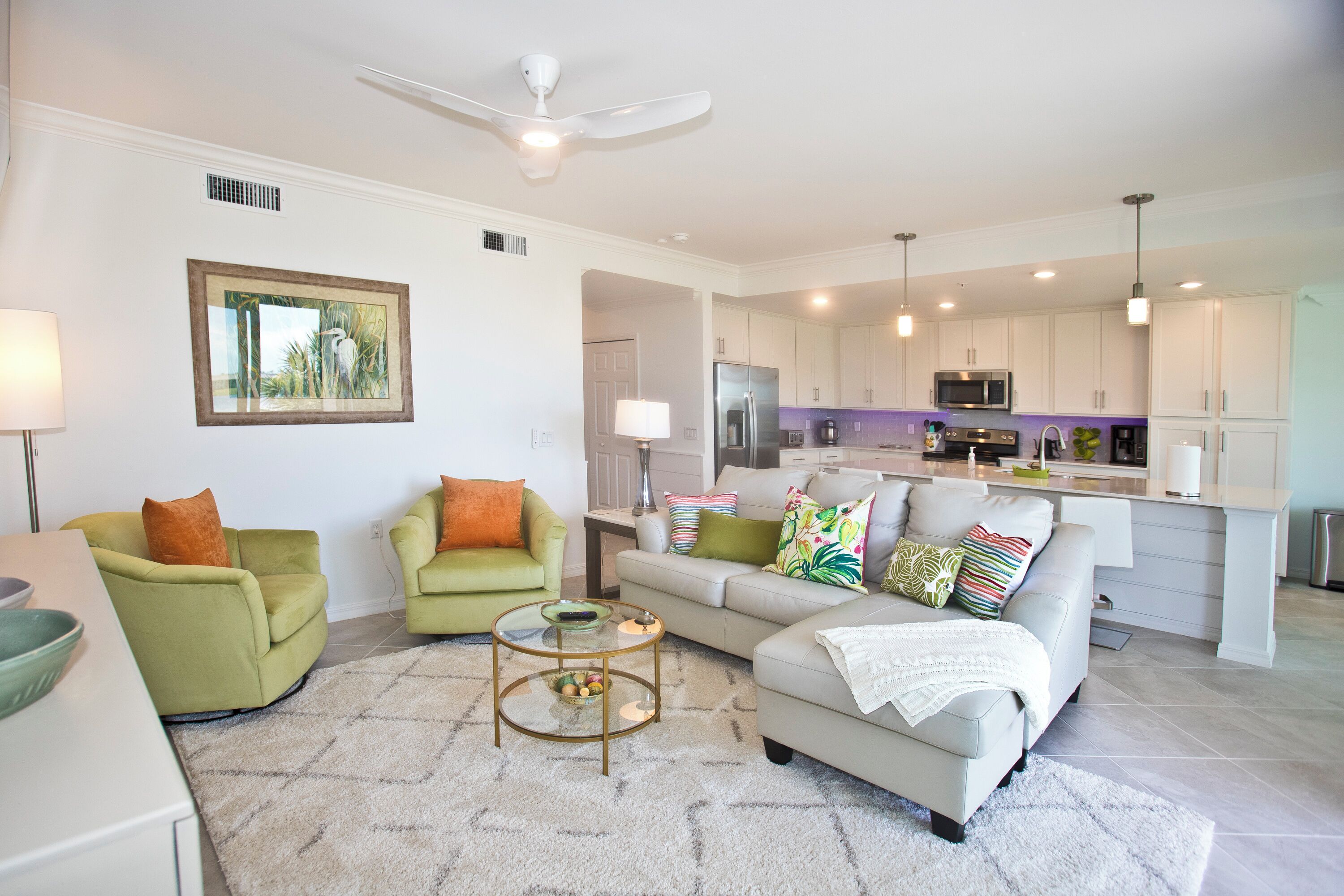 A modern living room with a sofa, chairs, and a coffee table. A kitchen is visible in the background with appliances and a dining area.