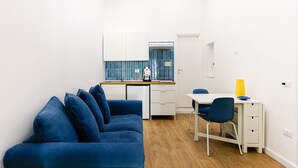 Ein kompakter Wohnbereich mit blauem Sofa, einem kleinen Esstisch mit Stühlen, einer Küchenzeile und einer weißen Tür.