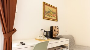 A hotel room with a white desk, a green chair, a bed with a striped blanket, a coffee maker, a remote control, and a framed fruit painting on the wall. - Santa Patrizia a Chiaia Apartment - Terra (Naples)