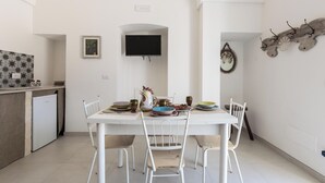 Comedor con una mesa y sillas blancas, un televisor montado en la pared y una cocina con un refrigerador y objetos decorativos.
