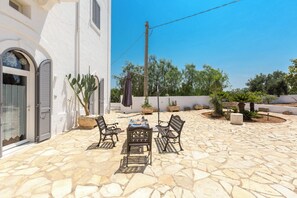 Un patio in pietra con tavolo e sedie, circondato da piante in vaso e un edificio bianco con finestre ad arco.