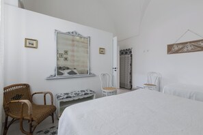 Una camera da letto con un letto grande, una sedia in vimini, una piccola panca, uno specchio e una porta che conduce a un'altra stanza.