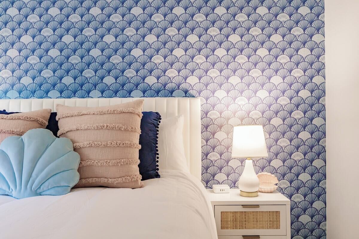 Une chambre à coucher avec un papier peint à motifs, un lit recouvert de plusieurs coussins et une table de chevet avec une lampe.
