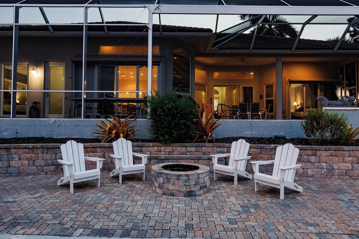 A patio area with four white Adirondack chairs arranged around a circular stone fire pit, set against a backdrop of a building with large windows and a covered outdoor area.