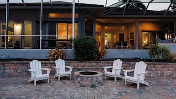 Eine Terrasse mit vier weißen Adirondack-Stühlen, die um eine runde Feuerstelle aus Stein angeordnet sind, mit Blick auf ein Gebäude mit großen Fenstern und einer überdachten Außenfläche.