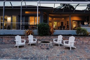 Un'area patio con quattro poltrone Adirondack bianche disposte attorno a un braciere in pietra circolare, con sullo sfondo un edificio con ampie finestre e una zona esterna coperta.