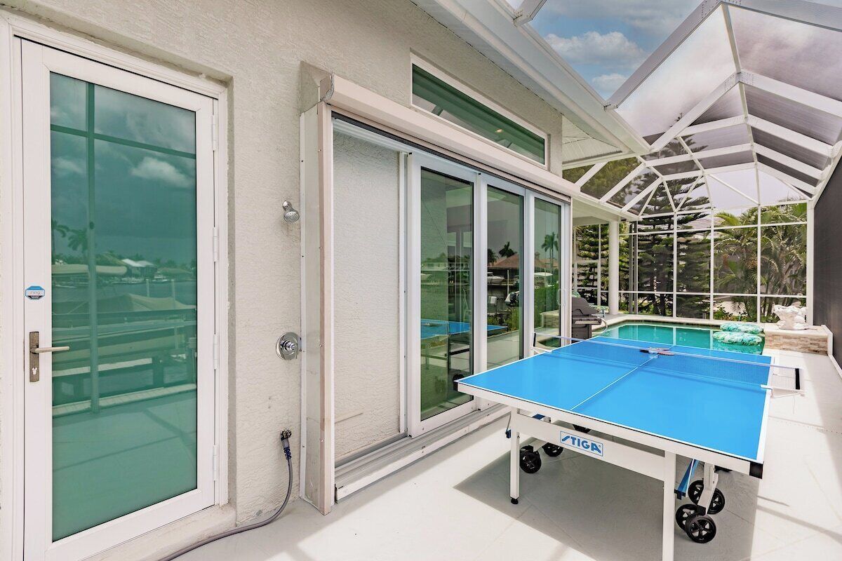 A ping pong table in a sunlit room with large windows and a pool in the background.