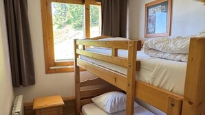 Ein Schlafsaal mit Etagenbetten, einem kleinen Holztisch, einem Fenster mit Vorhängen und einem gerahmten Bild an der Wand.
