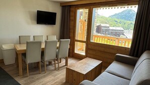 A room with a wooden dining table, four chairs, a sofa, a TV mounted on the wall, and a large window with a view of mountains. - Ski-in/ski-out & foot of the slopes, 4th floor, view ski slopes, swimming pool, balcony, tv (La Plagne)