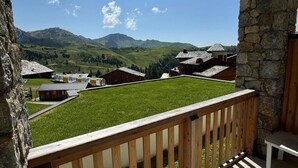 A balcony with a stone wall and wooden railing overlooking a village with traditional buildings and a mountainous landscape. - Ski-in/ski-out & foot of the slopes, 4th floor, view ski slopes, swimming pool, balcony, tv (La Plagne)