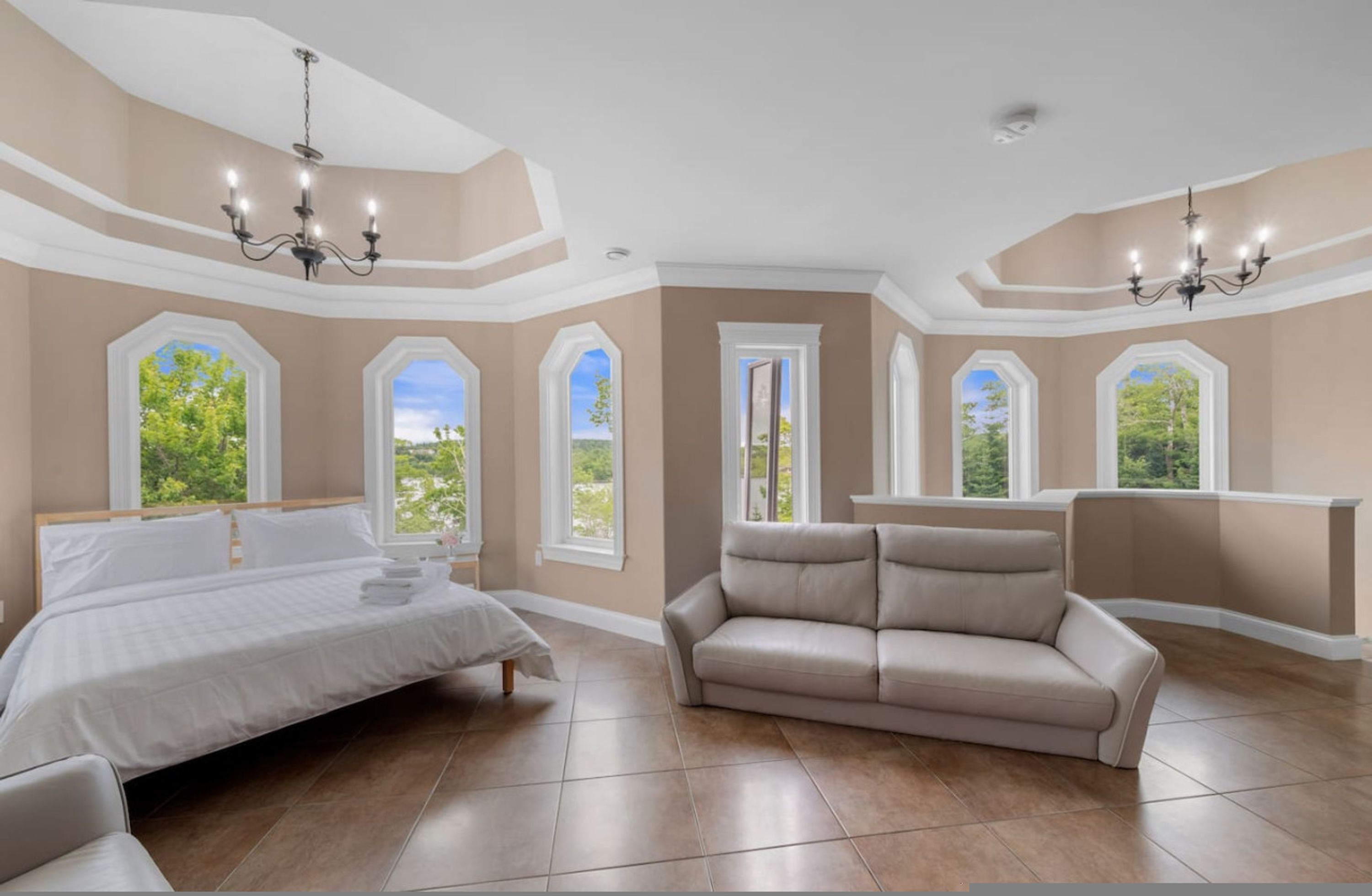 A spacious bedroom with a large bed, a beige sofa, and multiple windows offering a view of greenery.