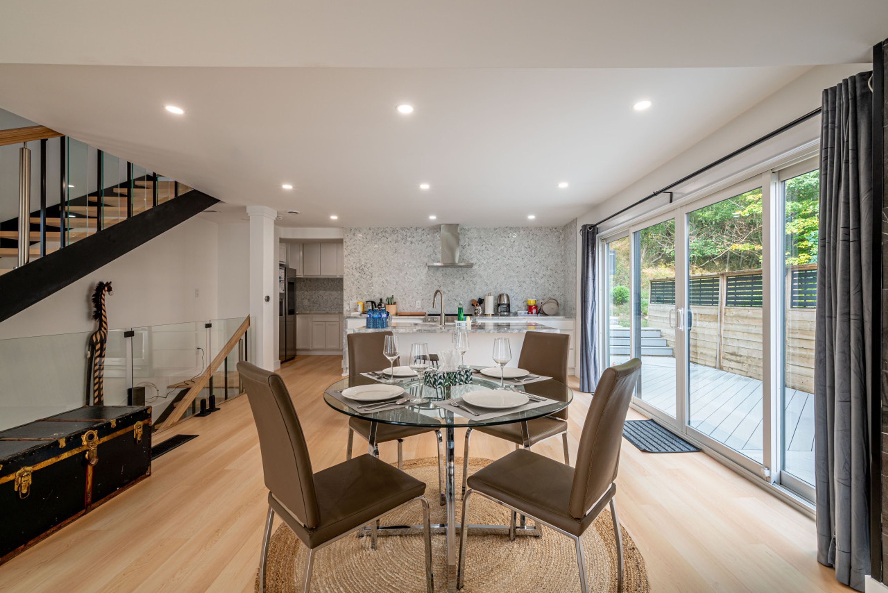 A modern dining area with a glass table, four chairs, and a kitchen in the background.