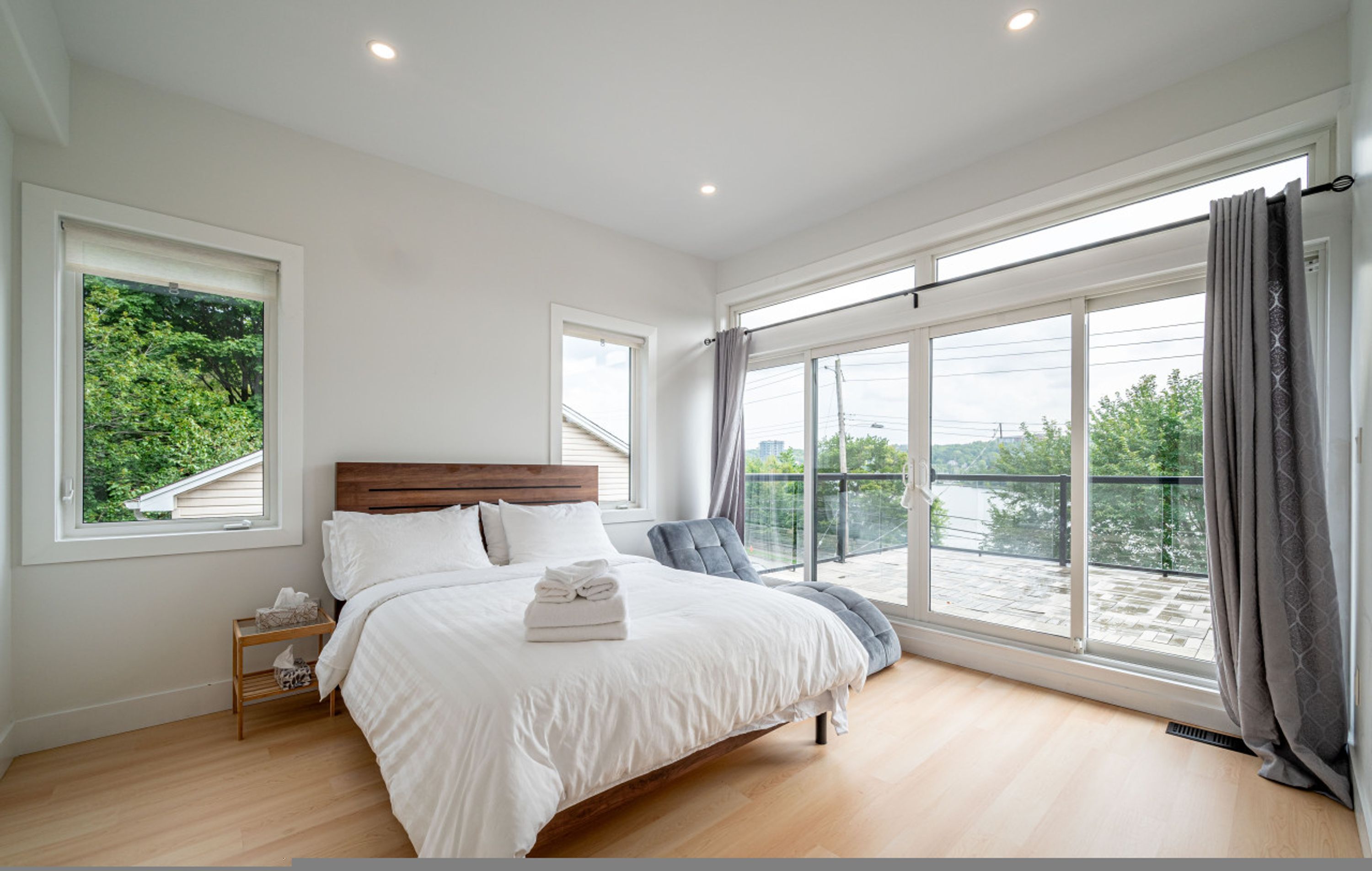 A bedroom with a large bed, two windows, a nightstand, and a view of greenery.