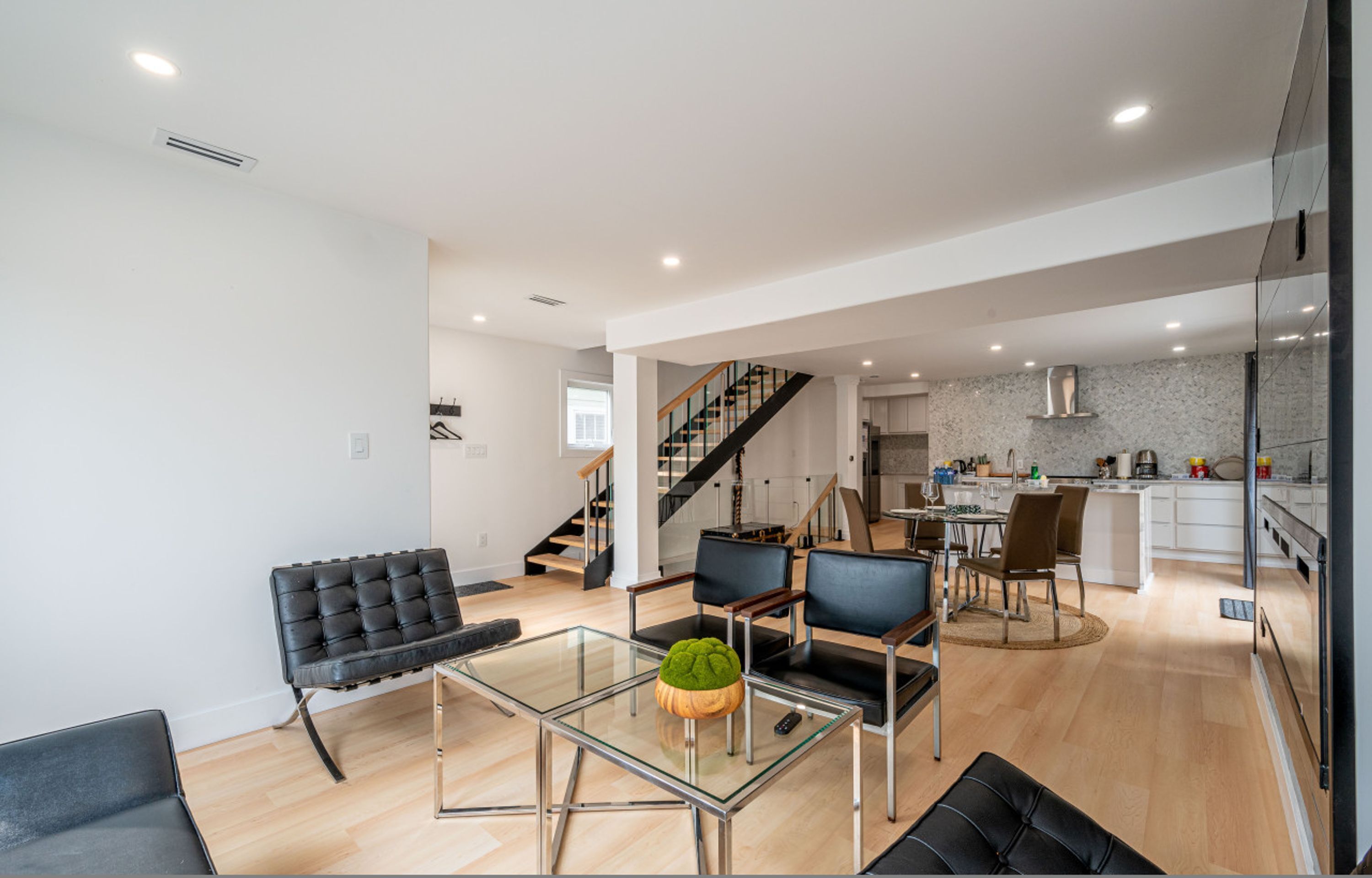 A modern open-plan living area with a glass dining table, black leather chairs, a staircase, and a kitchen in the background.