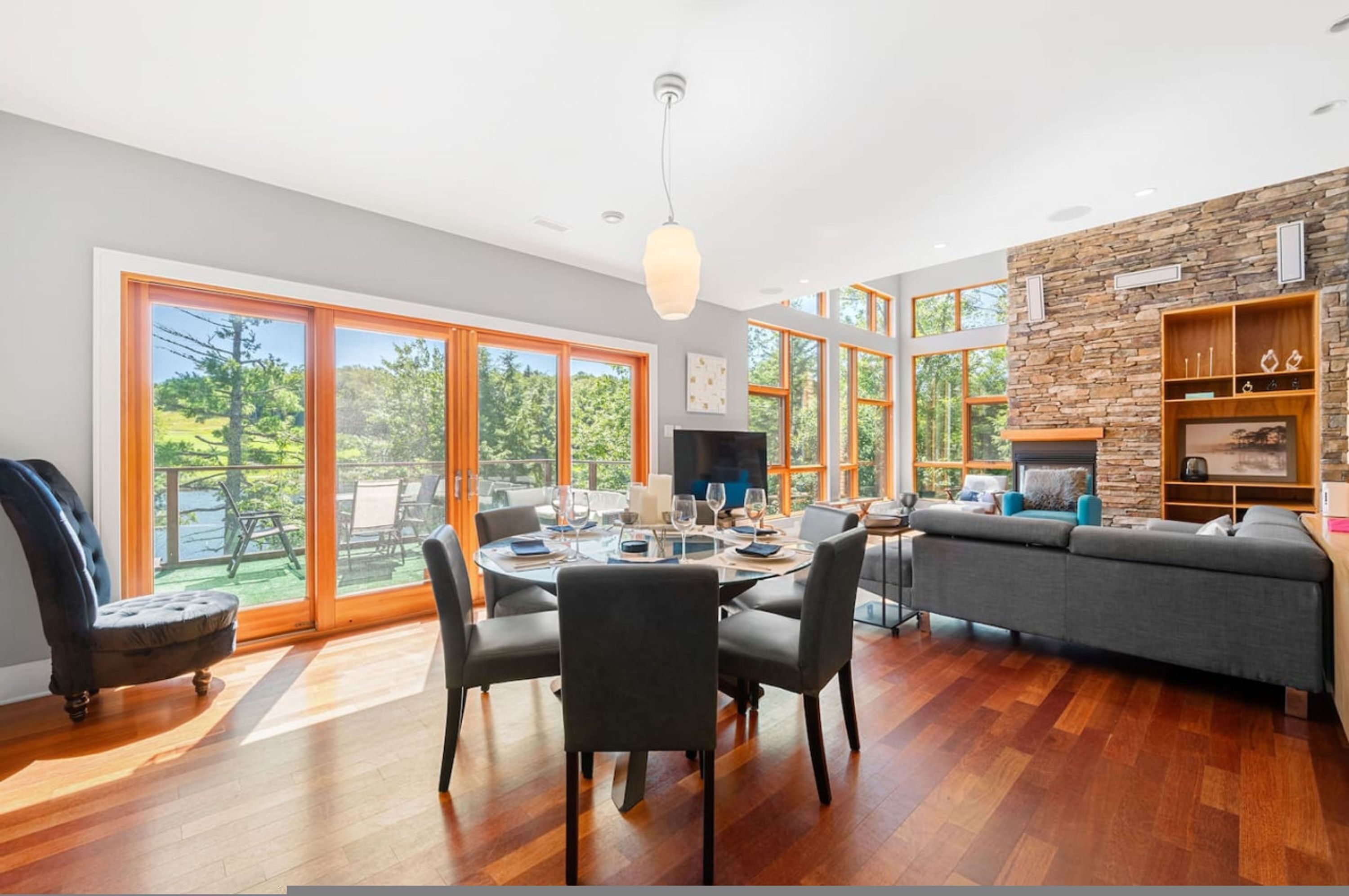 A spacious living area with a dining table, comfortable seating, a stone accent wall, and large windows offering a view of greenery.