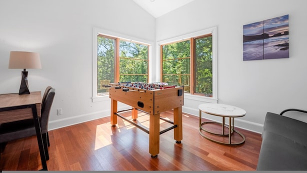 A room with a foosball table, a dining table with chairs, a round table, and large windows with a view of trees.