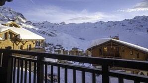 A wooden chalet with a snow-covered roof, a balcony, and a view of snow-covered mountains in the background.