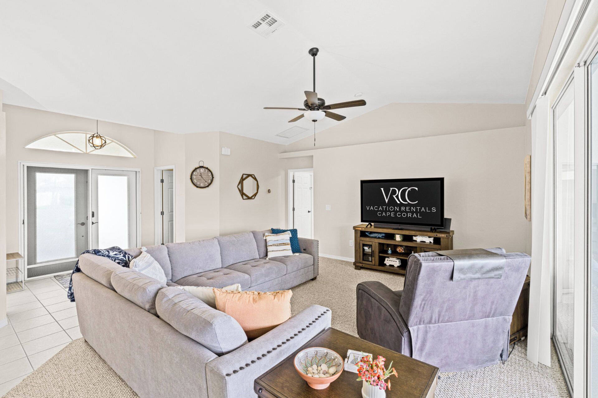 A modern living room with a grey sofa, a coffee table, and a TV. There is a ceiling fan, a clock, and a sign for Vacation Rentals Cape Coral.