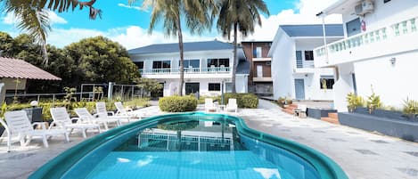 A swimming pool with lounge chairs, palm trees, and a building with balconies.