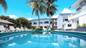 Una piscina con sillones de descanso, palmeras y un edificio de dos pisos con balcones.